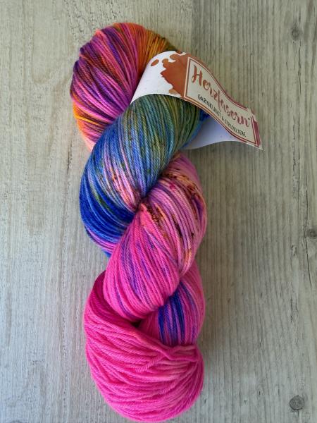 Handgefärbter Wollstrang in kräftigem Pink mit bunten Farbverläufen und Sprenkeln auf Herzfasern No 11 (Merino Superfine), fotografiert auf hellem Holzuntergrund mit Herzfasern-Banderole.